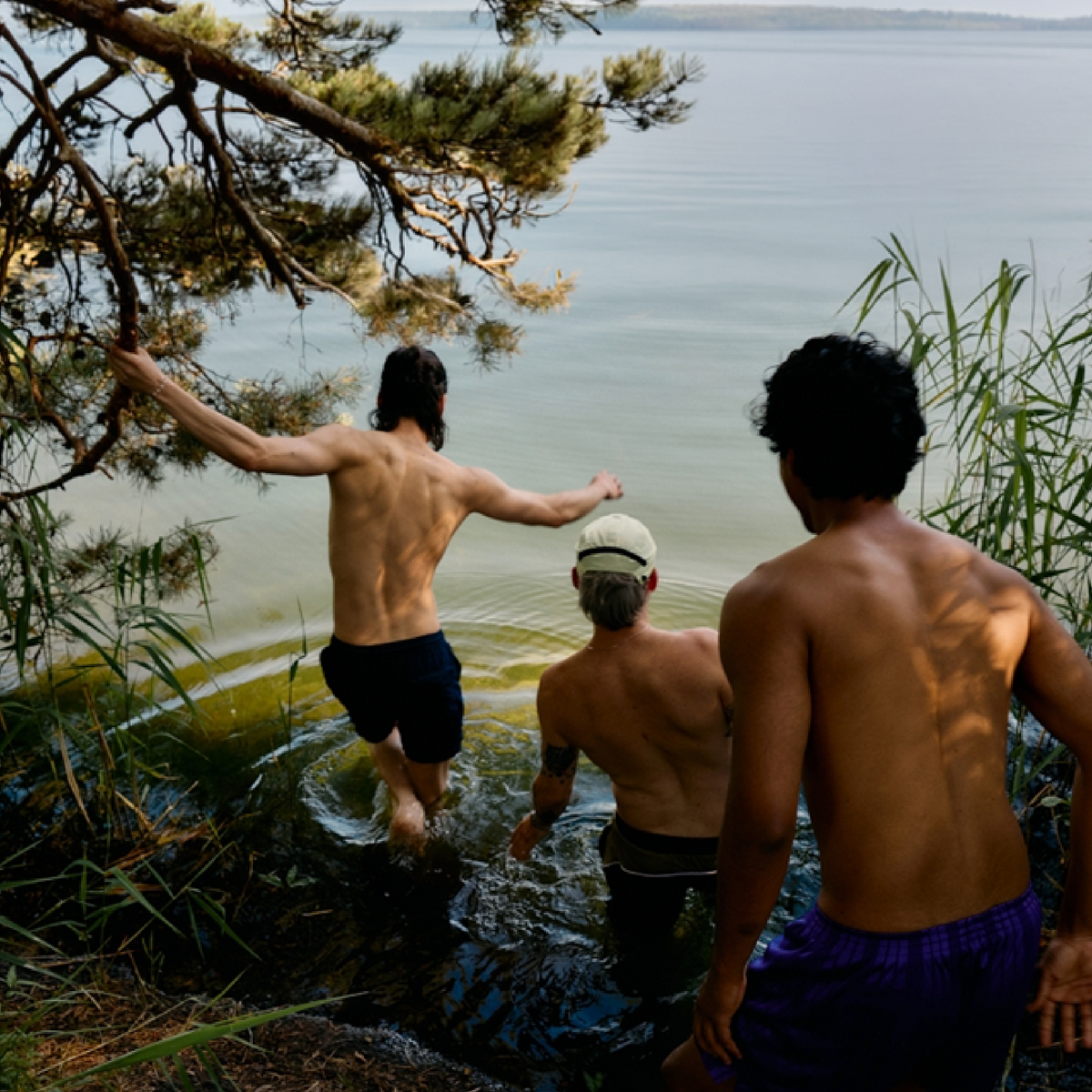 Three men standing by a lake, with one about to jump in.